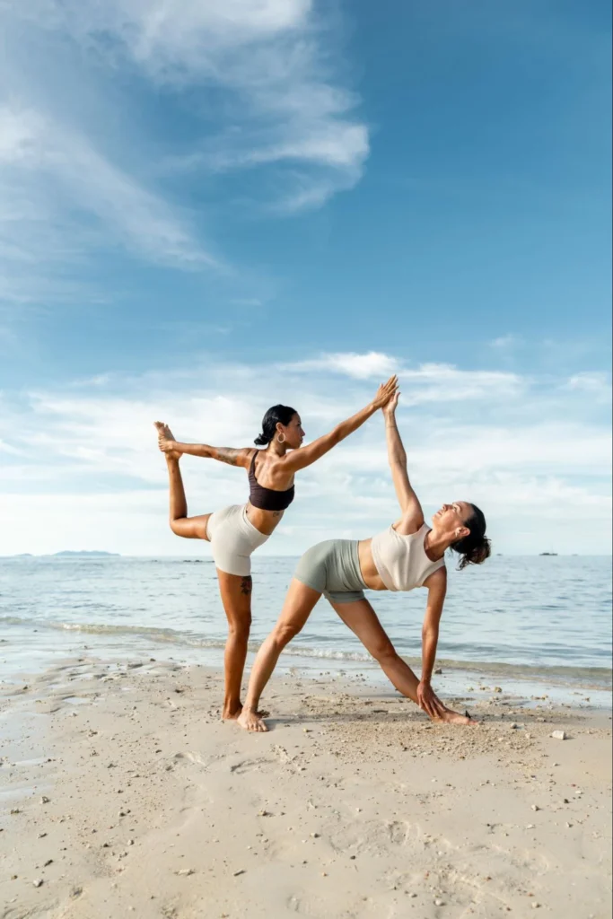 Two yoga students practicing partner standing poses on the beach during Vinyasa training