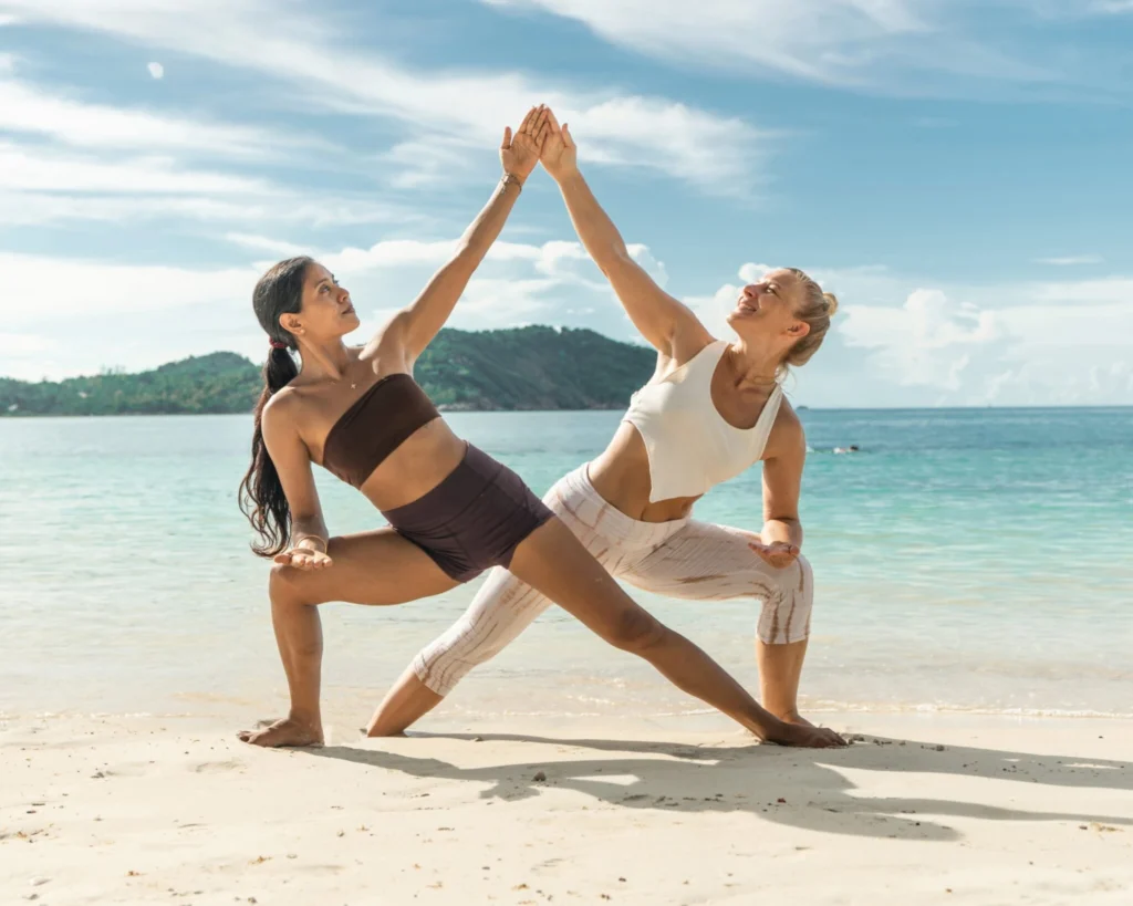 Two women practicing partner yoga on a tropical beach during yoga teacher training